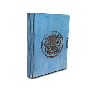 May include: A blue wooden journal with a laser-etched design featuring the word "Cthulhu" and an octopus-like creature. The journal has a clasp and a spine with vertical lines.