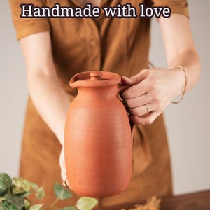 Puede incluir: Una jarra de terracota hecha a mano con tapa y asa. La jarra es de color marrón rojizo cálido y tiene una superficie ligeramente texturizada. El texto "Handmade with love" está en la parte superior de la imagen.