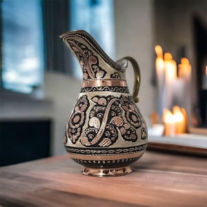 May include: A decorative copper pitcher with an ornate floral design. The pitcher features a curved spout, a small handle, and a base. The design includes black and copper accents. The pitcher is sitting on a wooden surface.