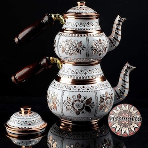 May include: White and copper-colored Turkish tea kettle with floral designs. The kettle has a stacked design with a lid, two spouts, and two wooden handles. The design includes copper bands and accents. A matching coaster is also visible.