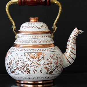 May include: White and copper-colored teapot with intricate floral and geometric designs. The teapot features a copper base, accents, and handle attachments. The handle is gold-colored with a wooden grip. The spout is white with copper stripes.