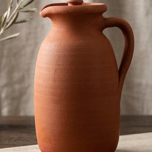 May include: A terracotta pitcher with a matte finish, featuring a rounded body, a curved handle on the right side, and a small spout. The pitcher includes a matching lid with a central knob, showcasing a natural reddish-brown clay texture.