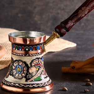 Könnte beinhalten: Eine kupferfarbene türkische Kaffeekanne mit einem dekorativen Blumendesign in Blau, Rot und Grün. Die Kanne hat einen langen, dunklen Holzgriff mit Blumenschnitzereien und einen Messinganschluss. Die Kanne ist zum Brühen von Kaffee bestimmt.