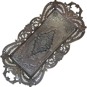 May include: An antique-style metal tray with an intricate, embossed design. The rectangular tray features a detailed pattern and a decorative, openwork border. The color is a muted bronze or silver, suggesting an aged appearance. Suitable for serving or display purposes.