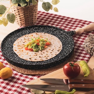 Puede incluir: Una plancha de cocina negra moteada con un mango de madera contiene una tortilla cubierta con guacamole y tomates en dados. Un paño a cuadros rojo y blanco y una tabla de cortar de madera con cuchillos y verduras están en el fondo.