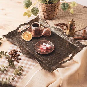 Puede incluir: Una bandeja de servicio ornamentada, de color bronce oscuro, con asas, que contiene una taza de café, delicias turcas y una taza y platillo decorativos de cobre. La bandeja está decorada con intrincados patrones y descansa sobre un mantel a cuadros.