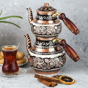 Puede incluir: Una tetera de cobre y negra con diseños florales y asas de madera. Un vaso de té, galletas, palitos de canela y rodajas de naranja secas están dispuestas cerca. La tetera tiene un diseño único de dos niveles.