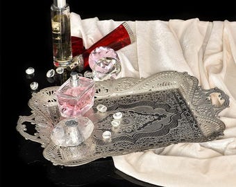 May include: A silver-colored, ornate tray holds perfume bottles, a red tube, and decorative crystals. A pink perfume bottle and a clear crystal stopper are in the center. The background includes a white cloth and a black surface.