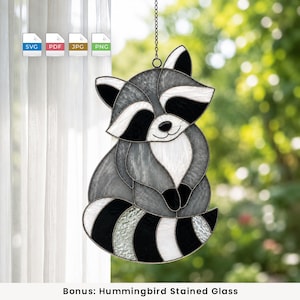 May include: A stained glass suncatcher shaped like a raccoon with grey, black, and white glass segments. The raccoon has a striped tail, pointed ears, and a dark mask around its eyes. It hangs from a metal chain against a window background.