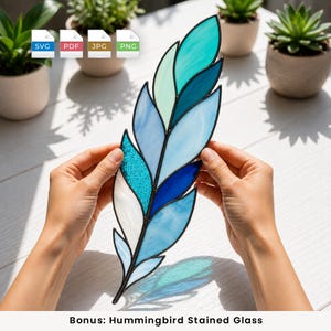 Pode incluir: Um ornamento de pena em vitral com um gradiente de segmentos de vidro azul, turquesa e branco unidos por linhas de solda de metal escuro. O design inclui peças de vidro texturizadas, translúcidas e opacas em vários tons de azul.