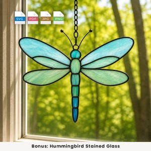May include: A dragonfly suncatcher made of stained glass with shades of light blue and green, featuring a black metal frame and a hanging chain. The wings are translucent, and the body displays a gradient from light green to deep blue.