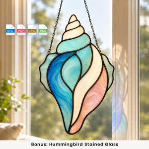 May include: A stained glass suncatcher shaped like a seashell, featuring segments of white, light blue, deep blue, and soft pink glass held together by dark metal solder lines. The piece hangs from a thin black metal chain against a window.