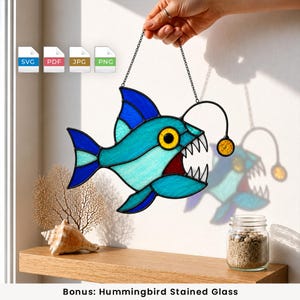 Può includere: Un acchiappasole in vetro colorato a forma di rana pescatrice nei toni del blu e del turchese, con un occhio giallo e un'esca luminosa. Il pesce ha denti bianchi affilati ed è appeso a una catena di metallo nero. Include icone di file SVG, PDF, JPG e PNG.