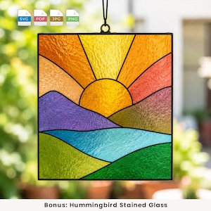 Puede incluir: Un atrapasoles de vidrieras cuadrado que presenta un paisaje con un sol amarillo brillante elevándose sobre colinas moradas y verdes, con un cuerpo de agua azul en primer plano. La pieza está enmarcada en metal negro y cuelga de un cordón negro.