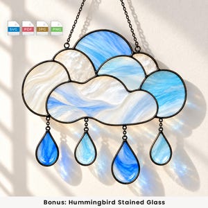 Puede incluir: Un atrapasoles de vidriera con forma de nube que presenta segmentos de vidrio blanco y azul con remolinos enmarcados en metal oscuro. Cuatro colgantes de vidrio azul en forma de lágrima cuelgan de la parte inferior con cadenas.