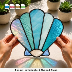May include: A seashell-shaped stained glass suncatcher featuring segments of iridescent, teal, light blue, and white glass held together by black metal solder lines. The piece displays a variety of textures and shades of blue and green.