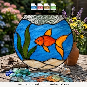 May include: A circular stained glass suncatcher featuring a bright orange goldfish swimming in blue water with green aquatic plants and a beige sandy bottom. The top section has clear textured glass. The design is framed in dark metal solder lines.