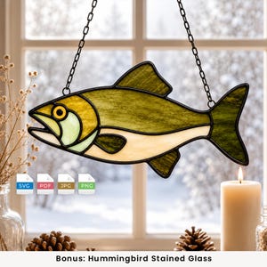 May include: A stained glass fish suncatcher featuring olive green, yellow, and cream-colored glass segments outlined in black metal. The fish is suspended by two black chains. Four small square icons labeled SVG, PDF, JPG, and PNG are visible.