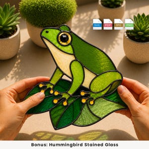 Pode incluir: Um ornamento de vitral representando um sapo com peças de vidro verde e amarelo unidas por linhas de solda de metal preto. O sapo está sentado sobre folhas verdes com pontas dos dedos amarelas e um olho grande, escuro e circular.