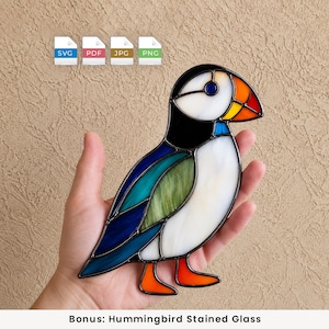 Könnte beinhalten: Ein Papageientaucher-Sonnenfänger aus Buntglas mit schwarzen, weißen, blauen, grünen und orangefarbenen Glasteilen, die durch dunkle Metalllötlinien verbunden sind. Oben im Bild befinden sich vier Dateiformat-Symbole: SVG, PDF, JPG und PNG.