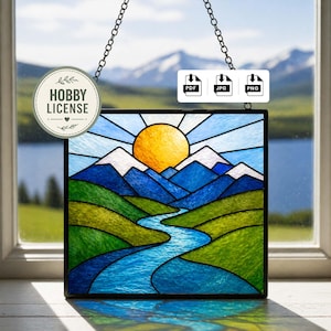 Op de afbeelding: Gebrandschilderd glaspaneel met een berglandschap met een gele zon, blauwe bergen en een rivier. Het paneel is zwart omlijst en hangt aan een ketting. Een sticker leest "HOBBY LICENSE".