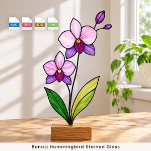 May include: A stained glass orchid ornament featuring two light purple and magenta flowers with yellow centres, two green leaves, and two small buds, all mounted on a rectangular wooden base. The design includes black metal lead lines.
