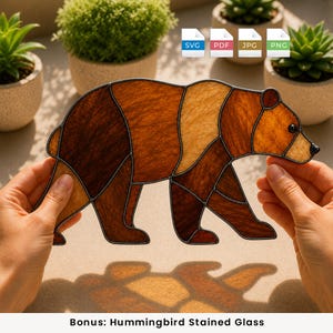 May include: A stained glass bear suncatcher featuring a segmented design with various shades of brown and tan glass pieces held together by dark metal solder lines. The bear is depicted in a walking profile with a small black eye and nose detail.