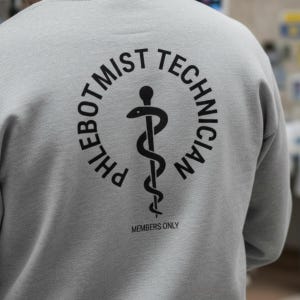 May include: A gray sweatshirt with the words "PHLEBOTOMIST TECHNICIAN" in a circular design, with a medical symbol in the center. Below the circle, the text reads "MEMBERS ONLY".