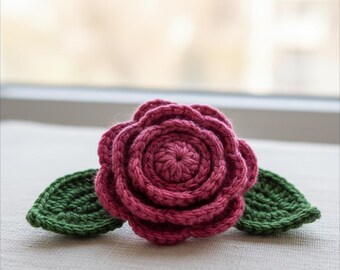 Puede incluir: Primer plano de una rosa tejida a ganchillo en rosa oscuro, con dos hojas verdes. La rosa presenta un intrincado diseño en espiral. Las hojas son de un verde contrastante. Probablemente sea una artesanía decorativa.