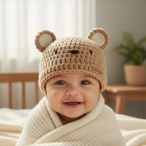 Könnte beinhalten: Ein lächelndes Baby trägt einen gehäkelten Bärenhut in Beige mit cremefarbenen Innenohren und einer schwarzen Nase und Augen. Das Baby ist in eine cremefarbene Decke gehüllt. Der Hut hat ein kleines braunes Etikett.
