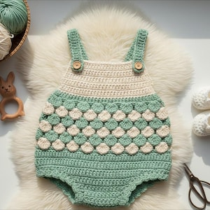 May include: A handcrafted crochet baby romper in mint green and cream. The romper has a scalloped pattern and wooden button fastenings. The image also shows a wooden bunny teether and a pair of crochet baby shoes.