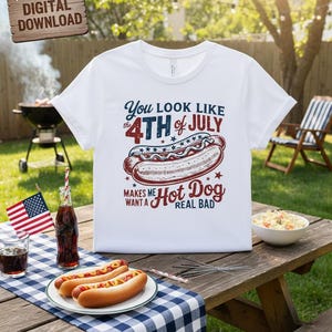 Puede incluir: Camiseta blanca con un gráfico que dice "You look like the 4th of July makes me want a hot dog real bad." La camiseta se muestra en una mesa de picnic con perritos calientes, bebidas y una bandera estadounidense.