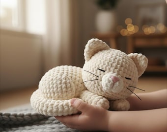 May include: A cream-colored crocheted cat plush toy with closed eyes and black whiskers. The cat is lying down, held in a person's hands. The cat's nose is pink. The plush toy is made of a soft, textured yarn.