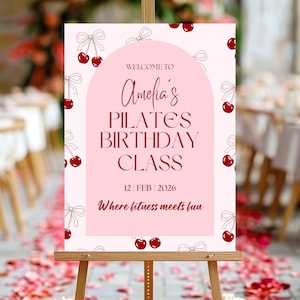 May include: A pink sign on a wooden easel with the text "Welcome to Amelia's Pilates Birthday Class" in burgundy script. The sign is decorated with cherries and bows. The date "12 FEB 2026" is also displayed.