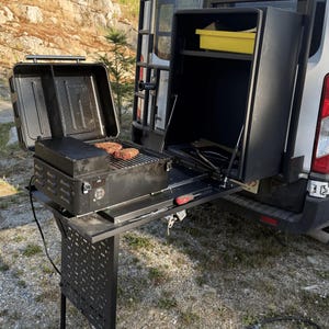 May include: A black portable barbecue and storage unit attached to the rear of a white van. The barbecue is open, with two steaks cooking on the grill. A black table extends from the unit, supported by legs. A yellow container is visible inside the storage unit.