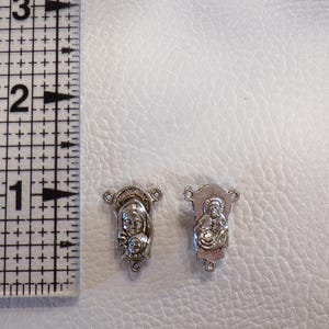 May include: Two small, silver-colored religious charms, each approximately 0.5 inches in length, are displayed on a white surface next to a ruler. The charms feature detailed depictions of religious figures. The ruler is marked with black lines and numbers.