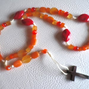 May include: A rosary with orange and red beads. The beads are various shapes, including oval and square. A small, dark cross hangs from the end of the rosary. The beads are strung on a white cord.