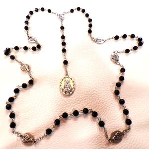 May include: A rosary necklace featuring black faceted beads and silver-toned metal accents. The necklace includes a crucifix pendant and several decorative medallions. The beads are strung on a delicate chain, creating a traditional religious accessory.