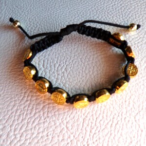 May include: A black macrame bracelet featuring gold-colored, coin-shaped beads. The bracelet has an adjustable sliding knot closure and gold-colored beads at the ends. The beads appear to have a religious symbol on them.