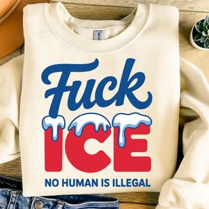 May include: A cream-coloured sweatshirt featuring the words "Fuck ICE" in blue script and red block letters, with white detailing. Beneath, the text "NO HUMAN IS ILLEGAL" is printed in navy blue.