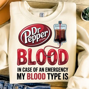 May include: Cream-colored sweatshirt with a humorous graphic. The design features the Dr Pepper logo, the word "BLOOD" in large red letters, and the phrase "IN CASE OF AN EMERGENCY MY BLOOD TYPE IS". A graphic of a blood bag is also included.