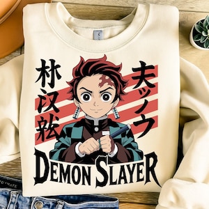 May include: Cream-colored sweatshirt featuring a graphic of an anime character with red hair and a scar. The design includes Japanese text and the words "Demon Slayer" in a bold font. The character is holding a sword.