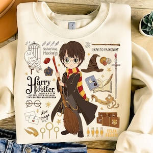 May include: Cream-colored sweatshirt featuring a cartoon illustration of Harry Potter holding a broom. The design includes an owl, a sorting hat, a wand, and the text "Harry Potter". Additional text includes "Showywe Hoobrig" and "Expvi to Paikonum".