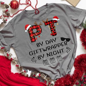 May include: A grey t-shirt featuring the text "PT by day, Giftwrapper by night." The letters "PT" are designed with a red and black plaid pattern, Christmas lights, and Santa hats. Sunglasses and gift box graphics are also included.
