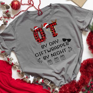 May include: A grey t-shirt with the text "OT by day, Giftwrapper by night." The letters "OT" are decorated with a red and black plaid pattern and Christmas lights. The shirt is surrounded by Christmas decorations.