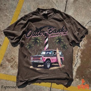 May include: Espresso-colored t-shirt featuring a graphic of a pink vintage SUV with a surfboard, palm trees, and a lighthouse. The text "Outer Banks Summer Feels" is arched above the scene. The shirt has a vintage, worn look.