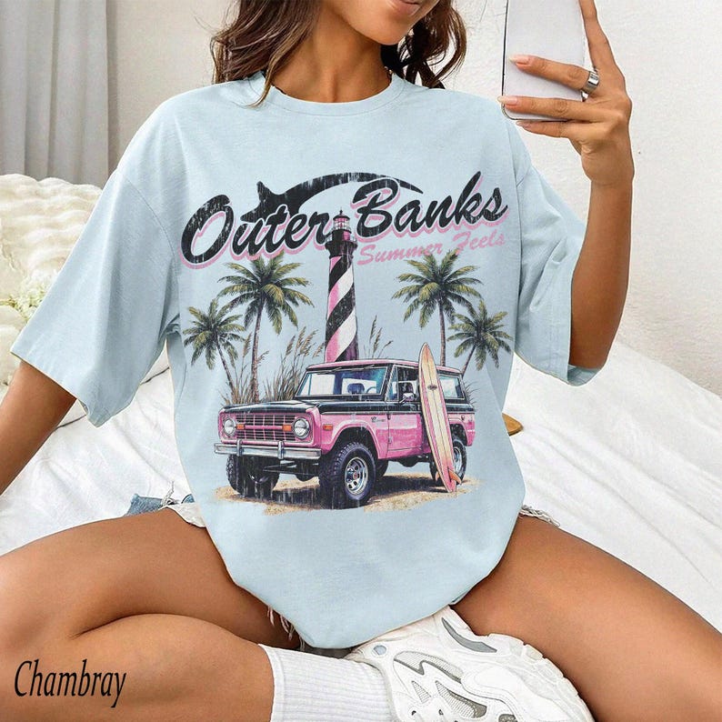 May include: Light blue t-shirt with an Outer Banks graphic. The design features a pink vintage SUV, a surfboard, palm trees, and a lighthouse. The text "Outer Banks Summer Feels" is printed above the image.