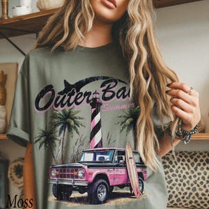 May include: Olive green t-shirt featuring a graphic of a pink vintage SUV, a lighthouse, palm trees, and the words "Outer Banks Summer". A surfboard leans against the SUV. The word "Moss" is printed at the bottom left.