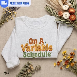 May include: A light gray crewneck sweatshirt featuring the text "On A Variable Schedule" printed in a retro-style font. The words "On A" and "Variable" are in orange with a yellow outline, while "Schedule" is in green with a yellow outline.