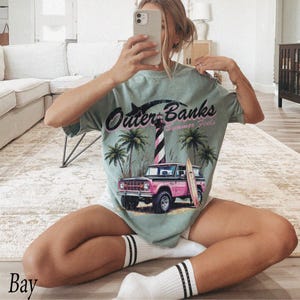 May include: A light green t-shirt with the words "Outer Banks Summer Feels" printed on the front. The graphic includes a pink and brown truck, a surfboard, palm trees, and a lighthouse. The shirt is a relaxed fit.